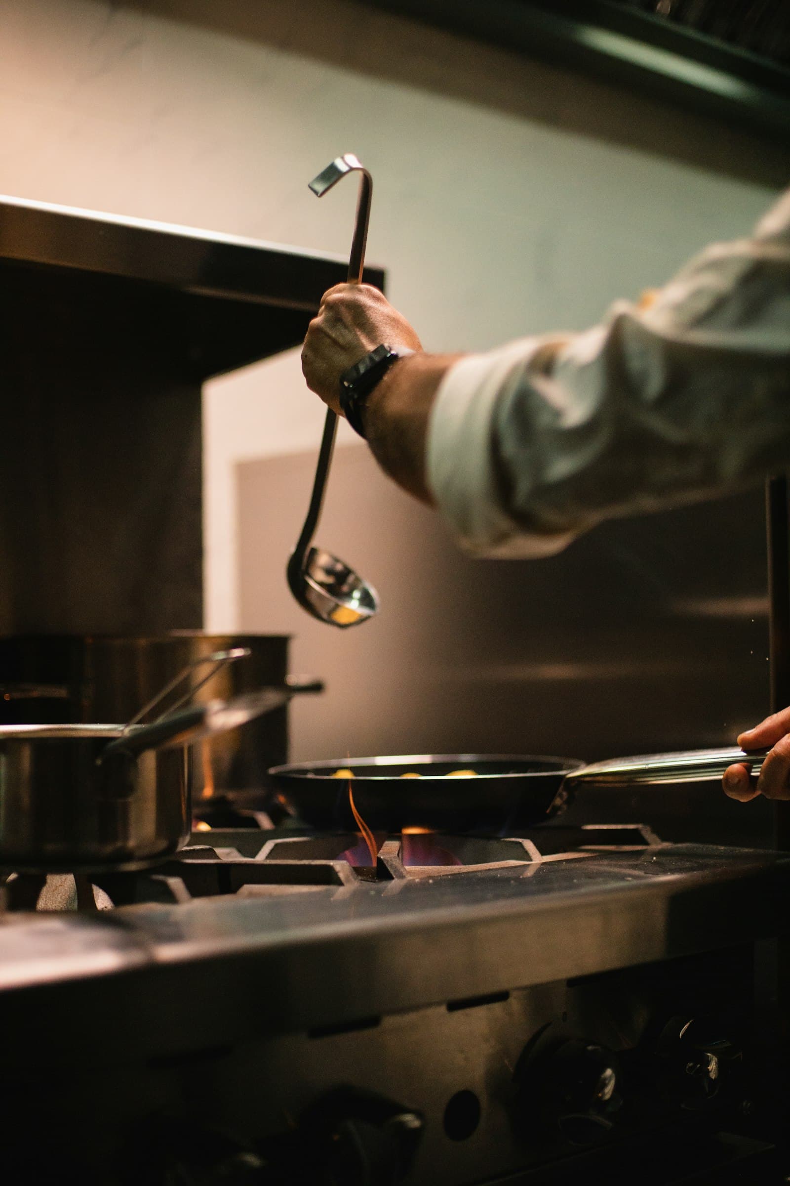 Hands cooking over a gas stove with a steel ladle, moody kitchen