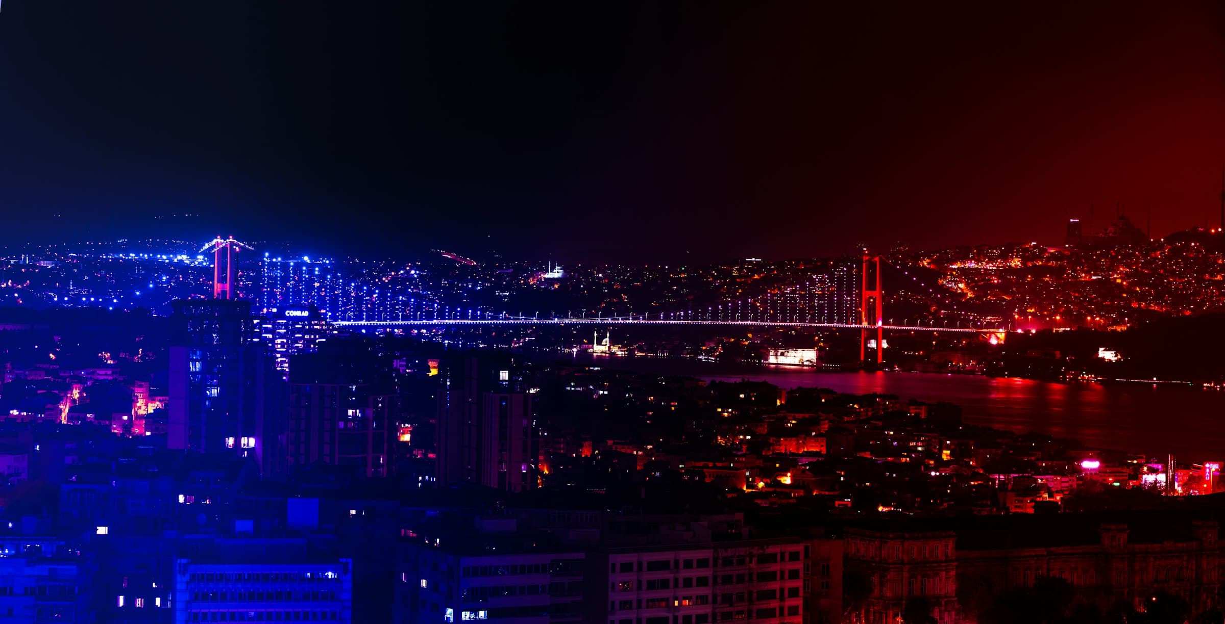 Aerial view of the Bosphorus Bridge lit blue and red over the strait at night